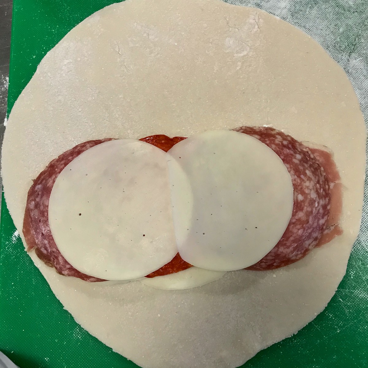 Image of a gluten free dough circle with a pile of lunch meat and slices of provolne cheese piled onto it 